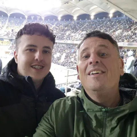 Mark Williams Two men. A younger man on the left has curly brown hair, a black jacket and braces. The older man on the right has short grey hair and is wearing a green jacket. They are both stood in the stands of a sports stadium and are smiling at the camera.