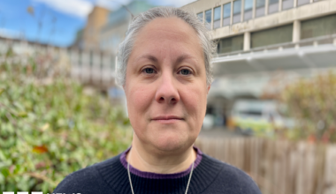 A headshot of Sian Coggle, who is standing with the Addenbrooke's Hospital building behind her. Sian has white hair tied back and is wearing a necklace and navy jumper.