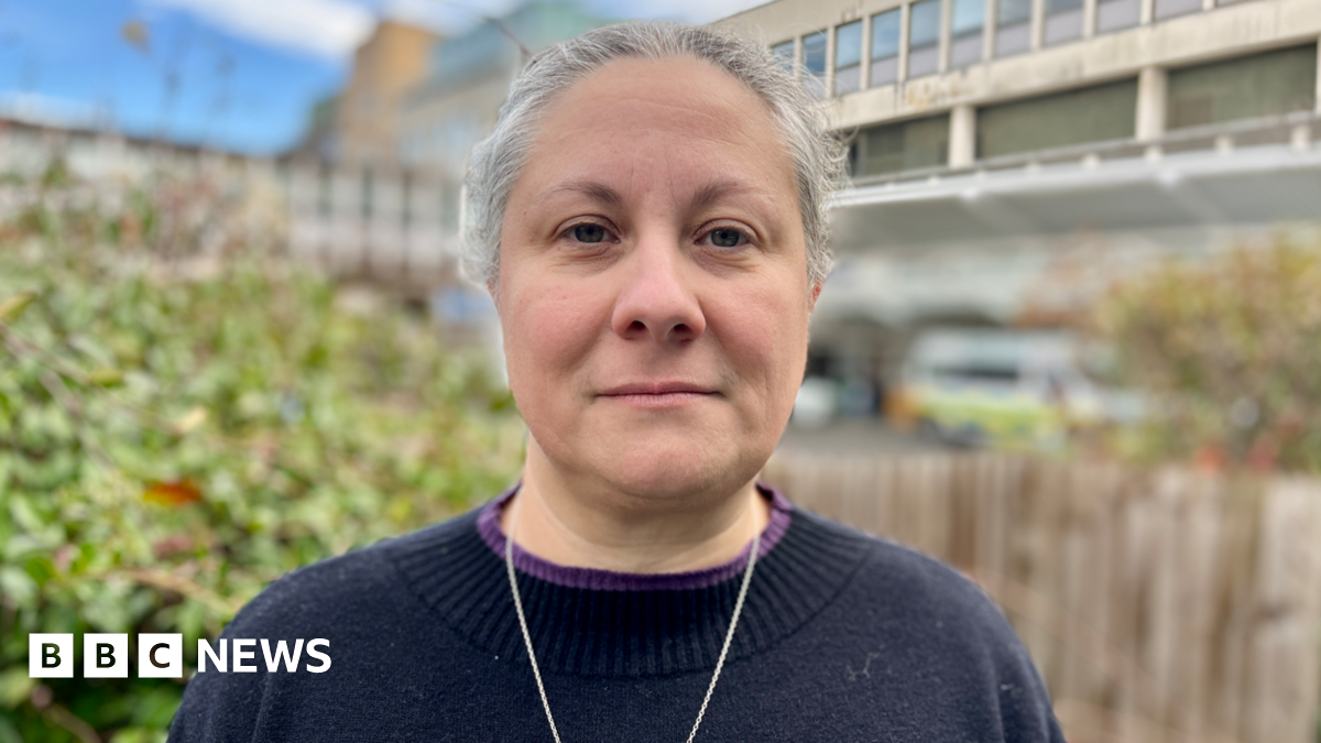 A headshot of Sian Coggle, who is standing with the Addenbrooke's Hospital building behind her. Sian has white hair tied back and is wearing a necklace and navy jumper.