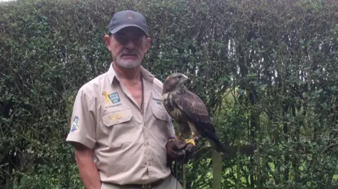 Mike Beresford Mike in a beige shirt with a dark cap on. He has a falcon on his left arm.