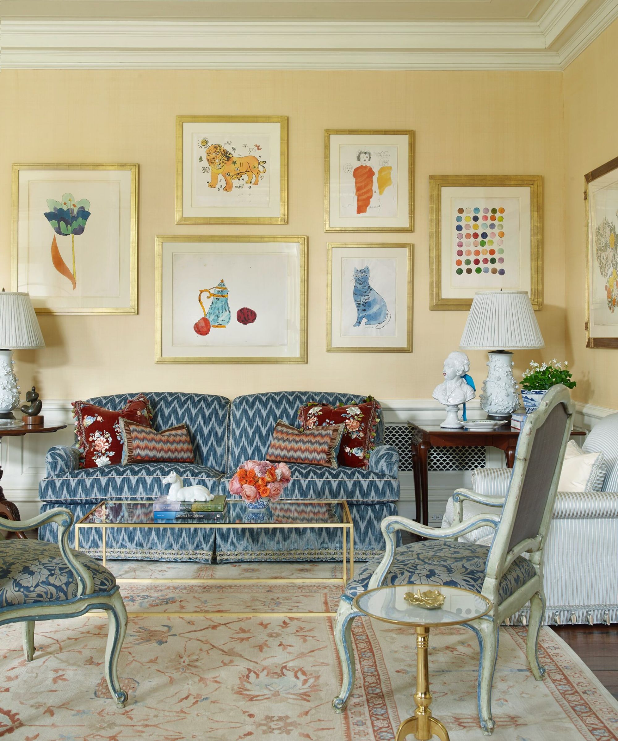A bright, elegant living room with pale yellow walls featuring a gallery wall of colorful whimsical art in gold frames. A blue and white ikat-patterned sofa sits behind a glass coffee table, flanked by large white ceramic lamps and vintage French-style armchairs