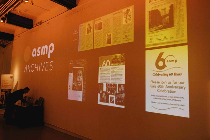 A wall displays "asmp ARCHIVES" alongside projected images of documents and flyers, including one inviting guests to a 60th anniversary celebration. A person stands at a table in the dimly lit room.