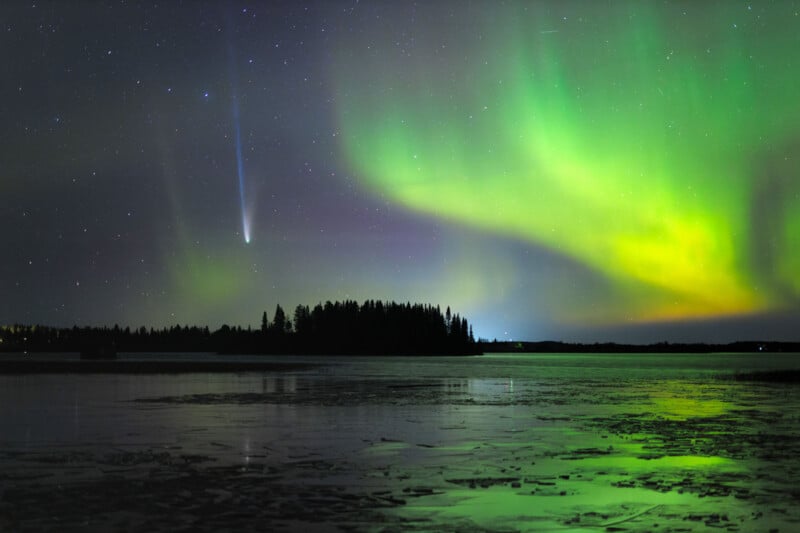 A comet streaks across a starry night sky above a forested island, with vibrant green and yellow auroras reflected on a calm, partially frozen lake in the foreground.