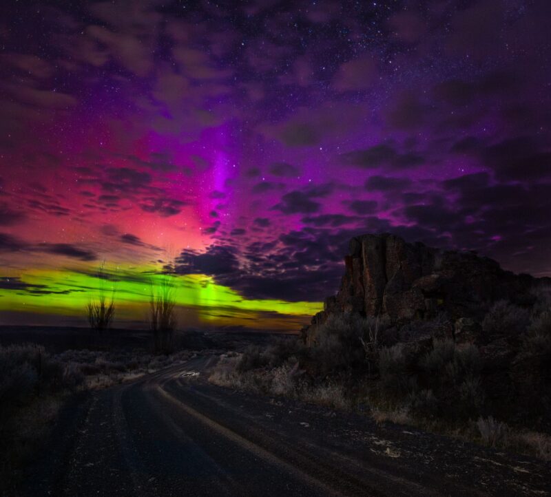 Forms of aurora: Rainbow colors of light near the horizon with dark clouds blocking patches and a dark rural landscape.