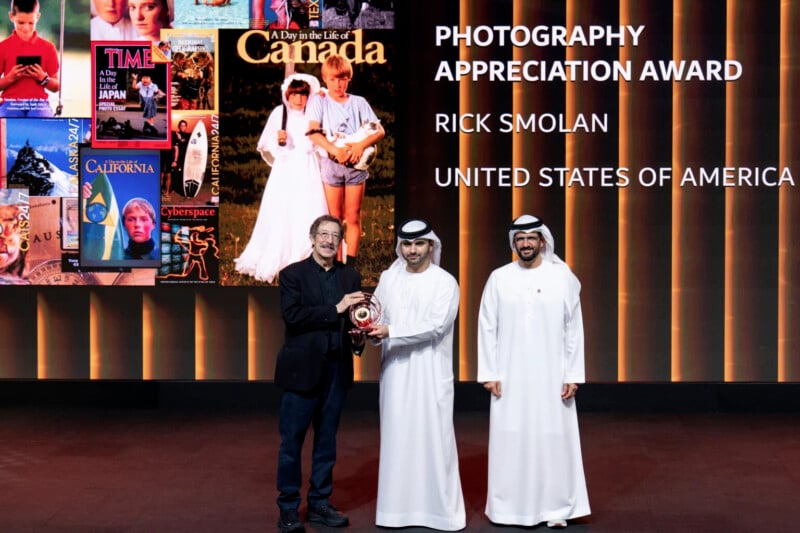 Three men stand on stage accepting the Photography Appreciation Award; behind them are magazine covers and text reading "PHOTOGRAPHY APPRECIATION AWARD, RICK SMOLAN, UNITED STATES OF AMERICA.