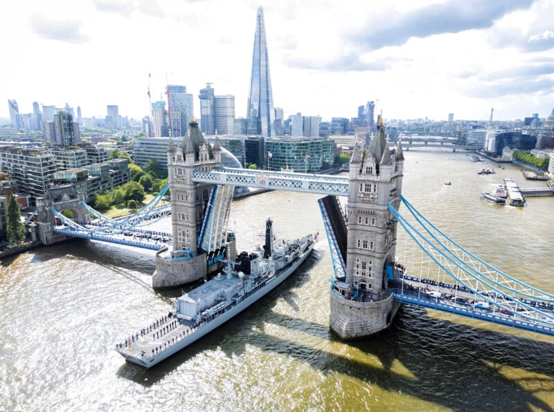 Tower Bridge in London is raised to allow a large naval ship to pass underneath. The city skyline, including The Shard, is visible in the background along the River Thames.