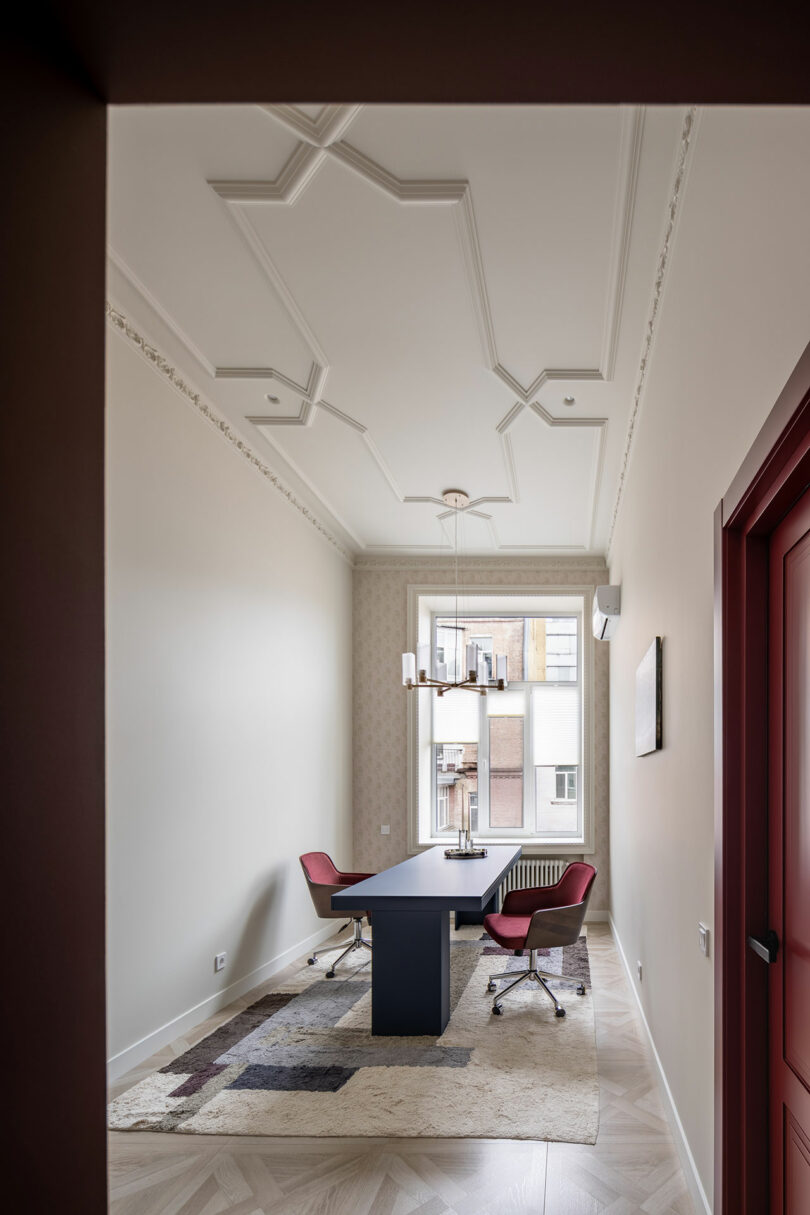 A modern office with a black table, two red chairs, a patterned rug, a chandelier, and a large window letting in natural light.