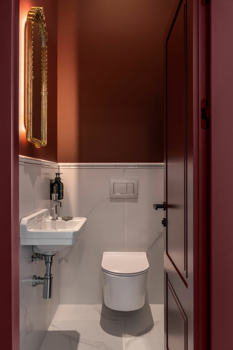 A small bathroom with white marble floor and walls, a wall-mounted toilet, a white sink, a soap dispenser, and a gold-framed mirror on a reddish-brown painted wall.