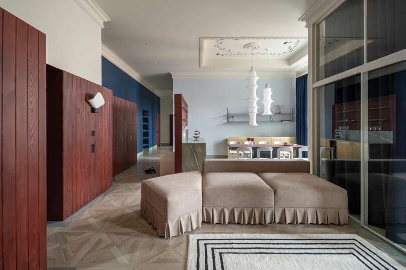 Modern living room with beige sectional sofa, geometric rug, blue and red accents, dining area in the background, and decorative ceiling moldings.