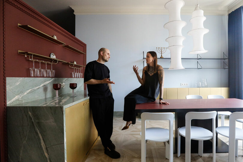 A man and woman talk in a modern kitchen; the woman sits on a counter while the man stands, both facing each other. The kitchen features red, gold, and marble accents.