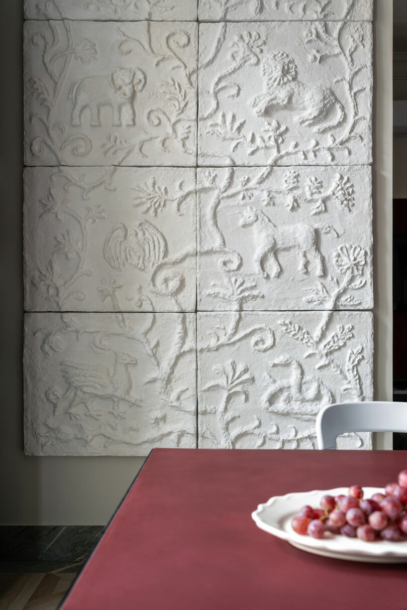 A textured white wall panel with animal and foliage carvings is seen behind a burgundy table with a white plate of grapes and a white chair in the foreground.
