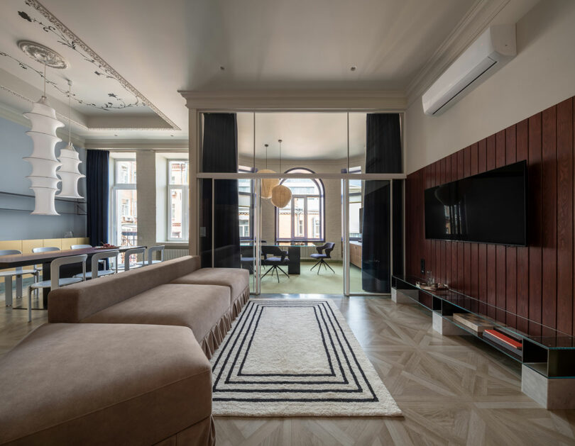 Modern living room with a brown sectional sofa, a geometric rug, a wall-mounted TV, wood paneling, and large glass doors opening to a bright dining area.