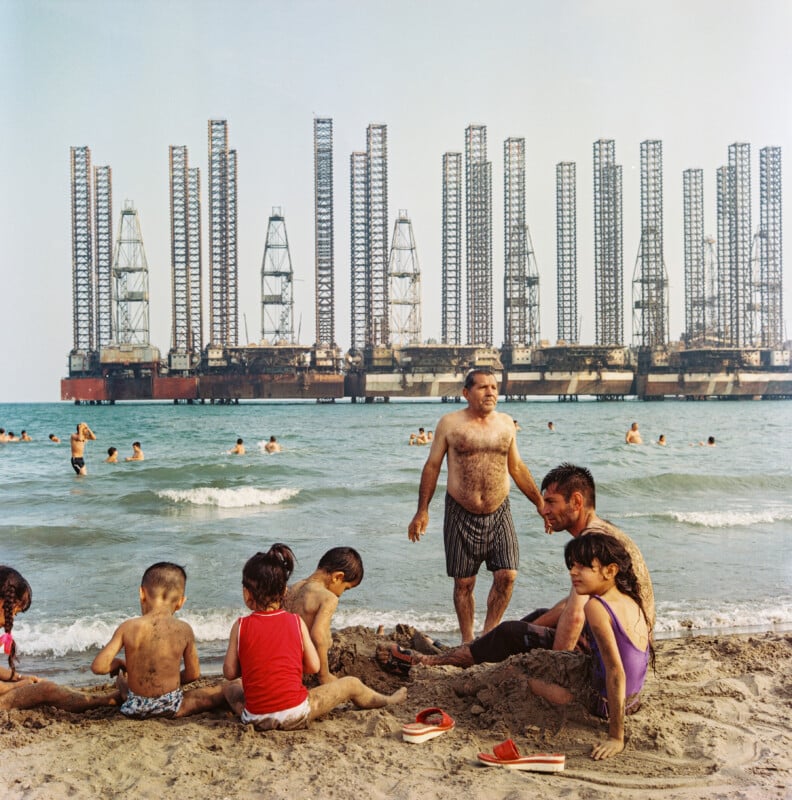 Children and adults play and relax on a sandy beach, while others swim in the sea. In the background, multiple large oil rigs are lined up along the horizon in the water.