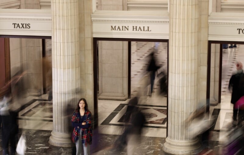 A young woman stands still in front of columns under a “MAIN HALL” sign, while blurred figures of people walk past her, creating a sense of motion in a busy indoor public space.