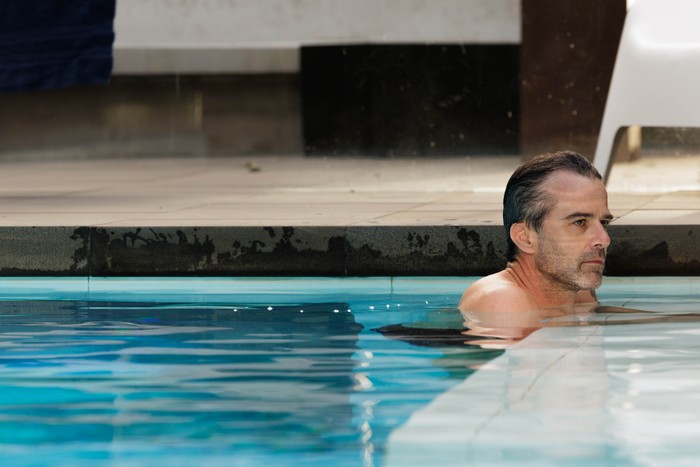 A man rests at the edge of an outdoor swimming pool with only his head and shoulders above the water, staring ahead with a serious expression.