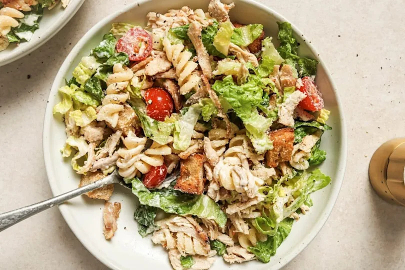 A fresh Caesar salad with chicken, croutons, tomatoes, and dressing, served in a large white bowl with a fork.