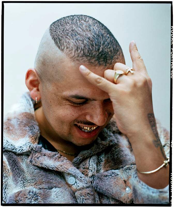 A smiling man wearing a furry jacket holds his hand to his forehead, showing rings and tattoos. He has short hair with shaved sides, gold teeth, and is looking down.