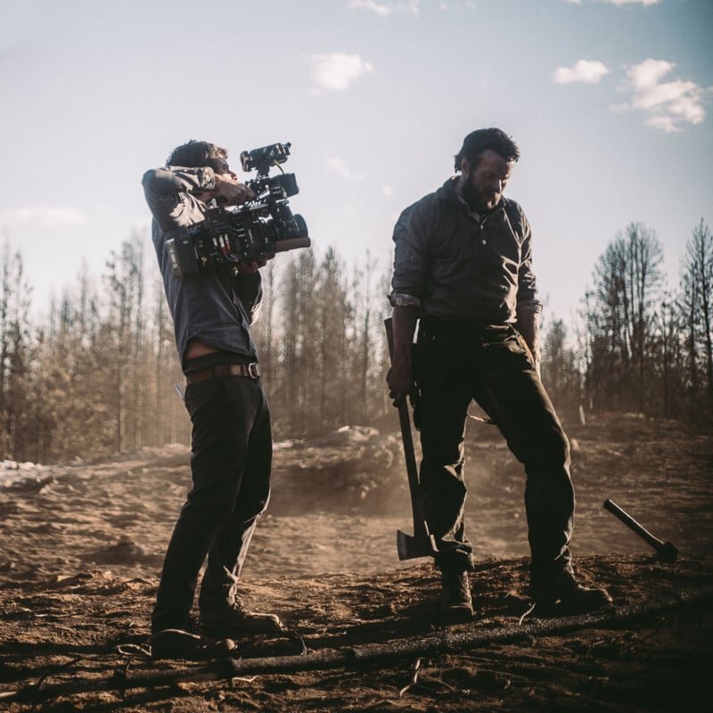 A filmmaker with a camera films a rugged man holding an axe outdoors on a dirt field, with bare trees and a cloudy sky in the background, evoking a dramatic, cinematic scene.