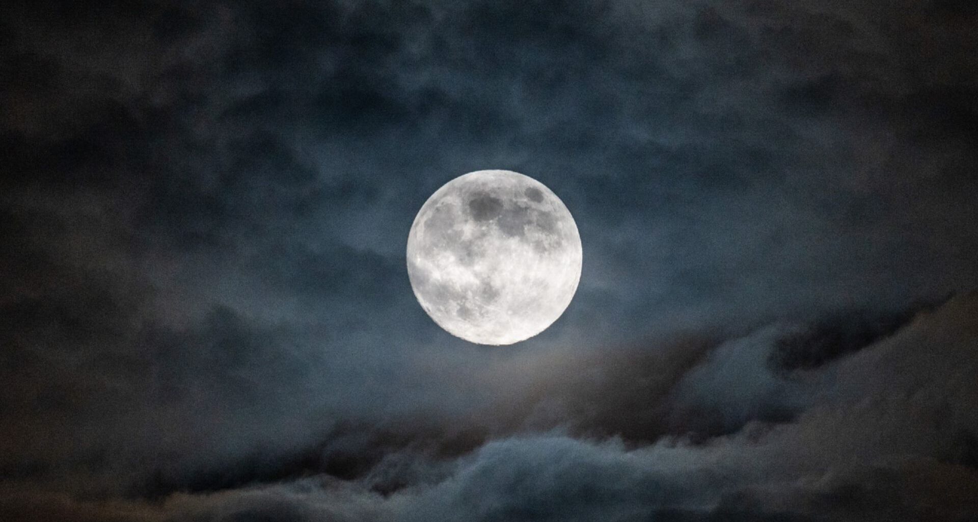 A full moon is pictured in a cloudy night sky, with the ancient lava plains known as lunar maria darkening its otherwise pearlescent surface.