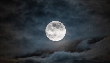 A full moon is pictured in a cloudy night sky, with the ancient lava plains known as lunar maria darkening its otherwise pearlescent surface.