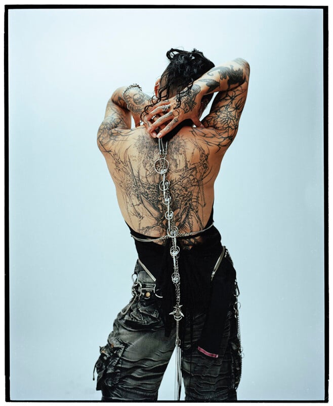 A person with extensive tattoos on their back and arms stands facing away, arms raised and hands clasped behind their head. They wear layered dark clothing and a long, decorative chain draped down their back.