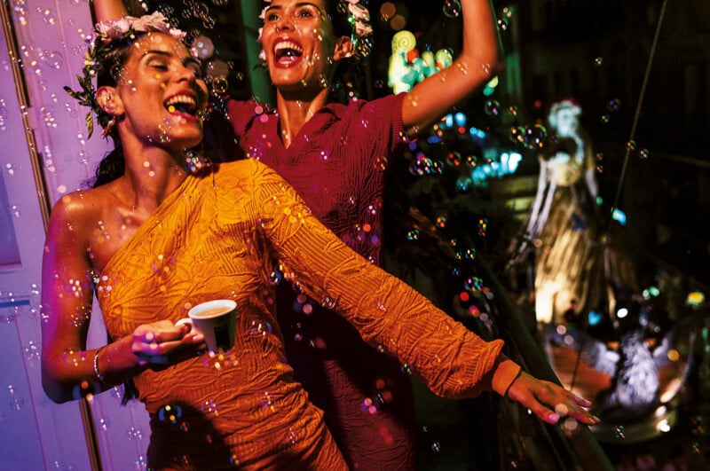 Two women in brightly colored dresses laugh and celebrate on a balcony at night, with one holding an espresso cup and the other raising her arms, surrounded by bubbles and city lights.