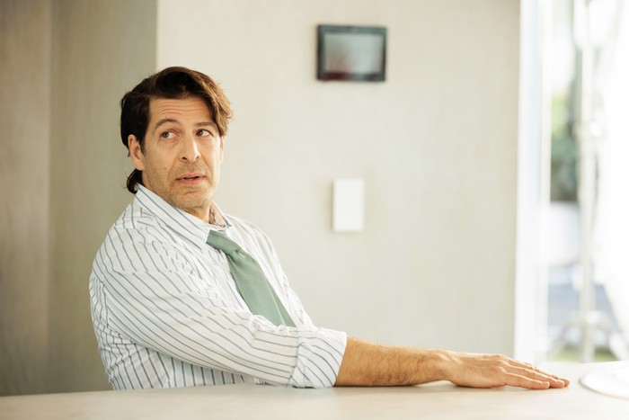 A man in a striped shirt with rolled-up sleeves and a loose green tie sits at a table, looking off to the side in a bright, minimalist room.