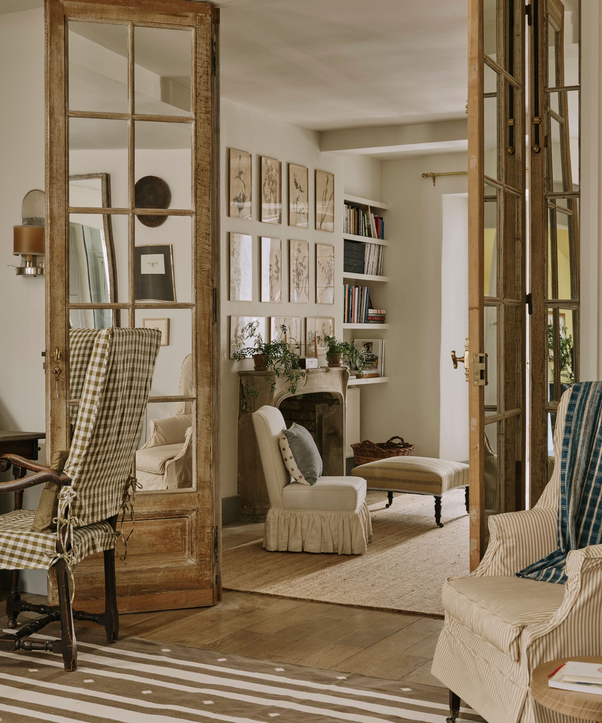 A view through weathered wood French doors into a serene, neutral-toned living space. The room features a stone fireplace, a gallery wall of botanical sketches, and cozy slipcovered furniture, including an armchair with a pleated skirt and a green-and-white gingham chair in the foreground