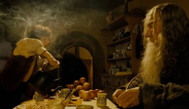 A young man pours tea from a kettle as an older bearded man sits at a wooden table with apples, mugs, and snacks in a cozy, warmly lit kitchen surrounded by shelves and dishes. Steam rises in the air.