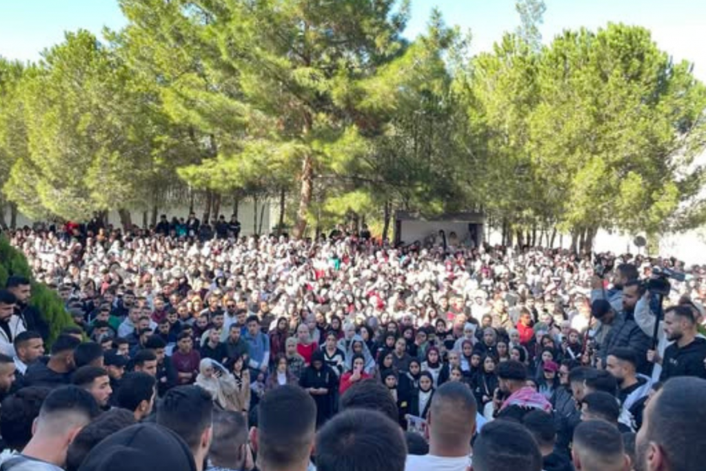 A very large crowd of young people gathered outdoors in a green, treed area. Many people are visible, packed together, facing a central point.