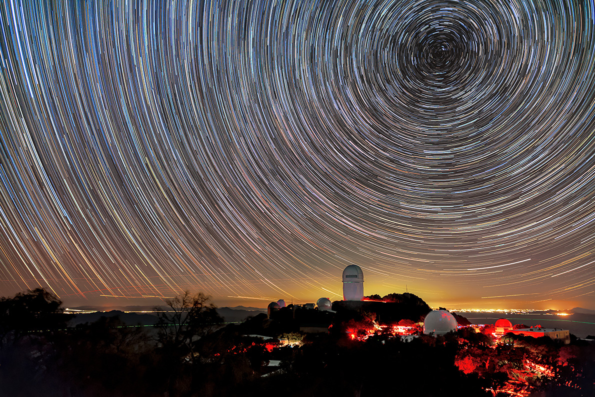 A series of circular star trails are seen in a purple night sky with an observatory below illuminated in red light.