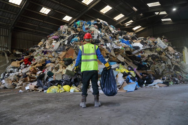 A man stands in a high-viz vest facing away from the camera in front of a pile of trash in a large hangar-like room