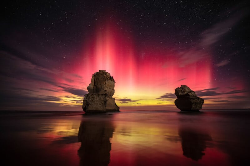Bright pink and yellow aurora lights illuminate the night sky above two large rock formations in the ocean, with starry sky and colorful reflections on the water's surface.