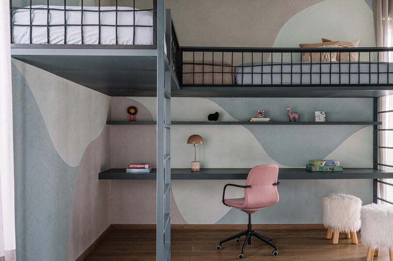 A modern loft-style kids' bedroom features two raised beds, a desk with a pink chair, open shelves with toys and books, and abstract wall designs.