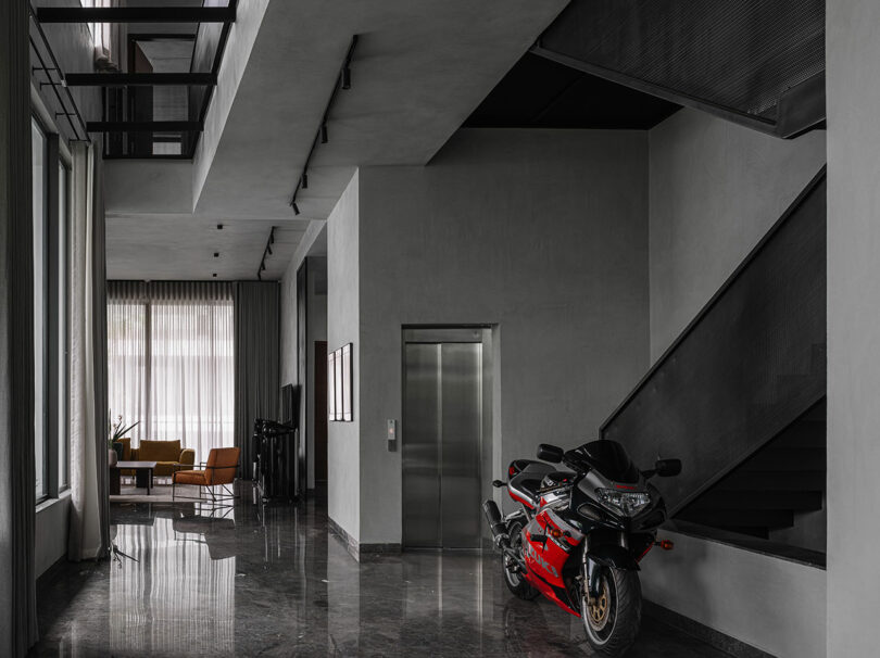 A red and black motorcycle is parked indoors near a staircase and an elevator in a modern, minimalist building with marble floors and large windows.