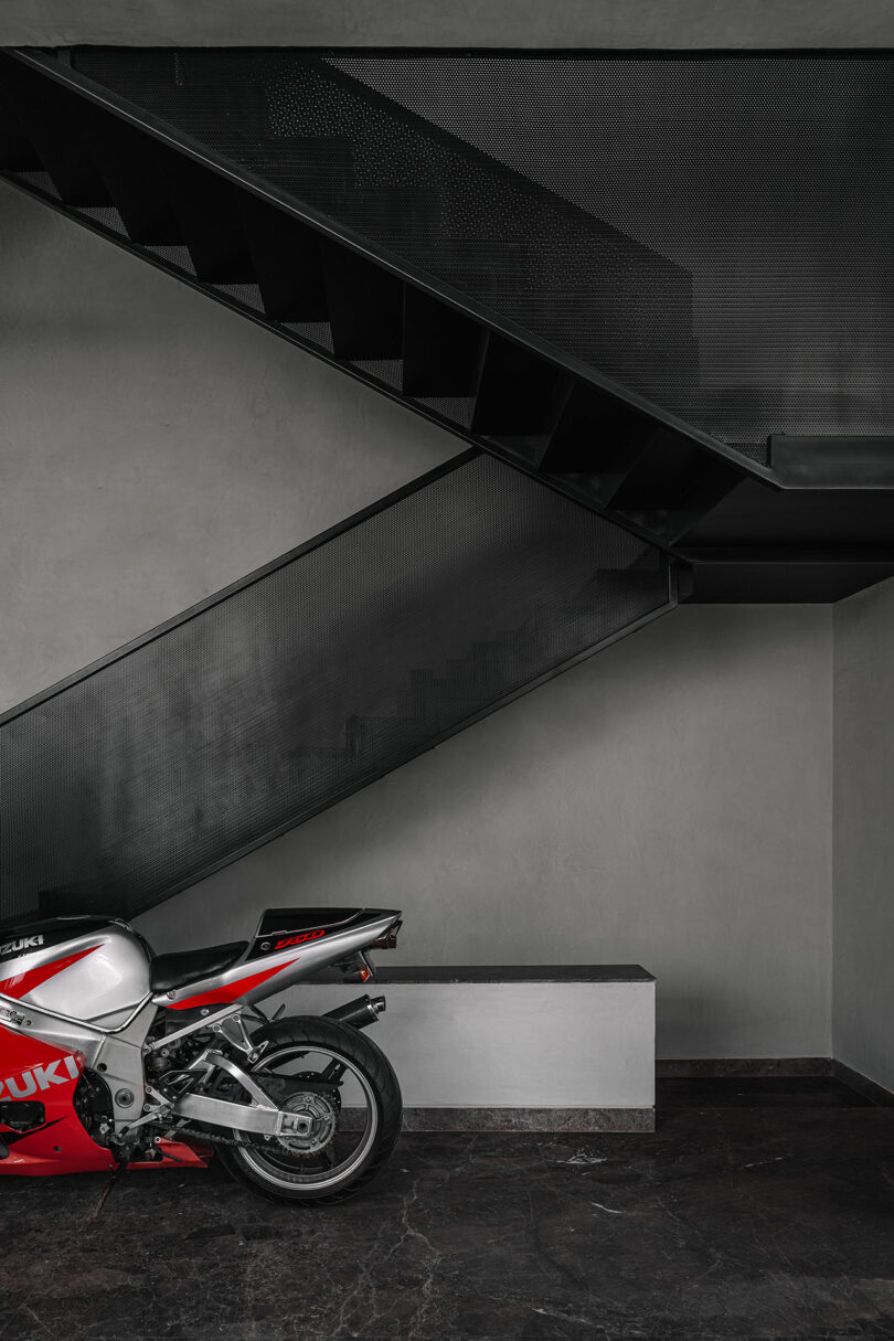 A red and white Suzuki motorcycle is parked indoors beneath a modern black metal staircase with a minimalist grey and black interior.