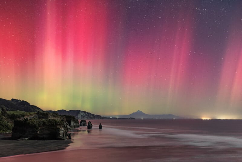 Colorful aurora lights in red, pink, and green hues illuminate the night sky above a coastal landscape with rocky cliffs, sea stacks, calm water, and distant mountains under a starry sky.