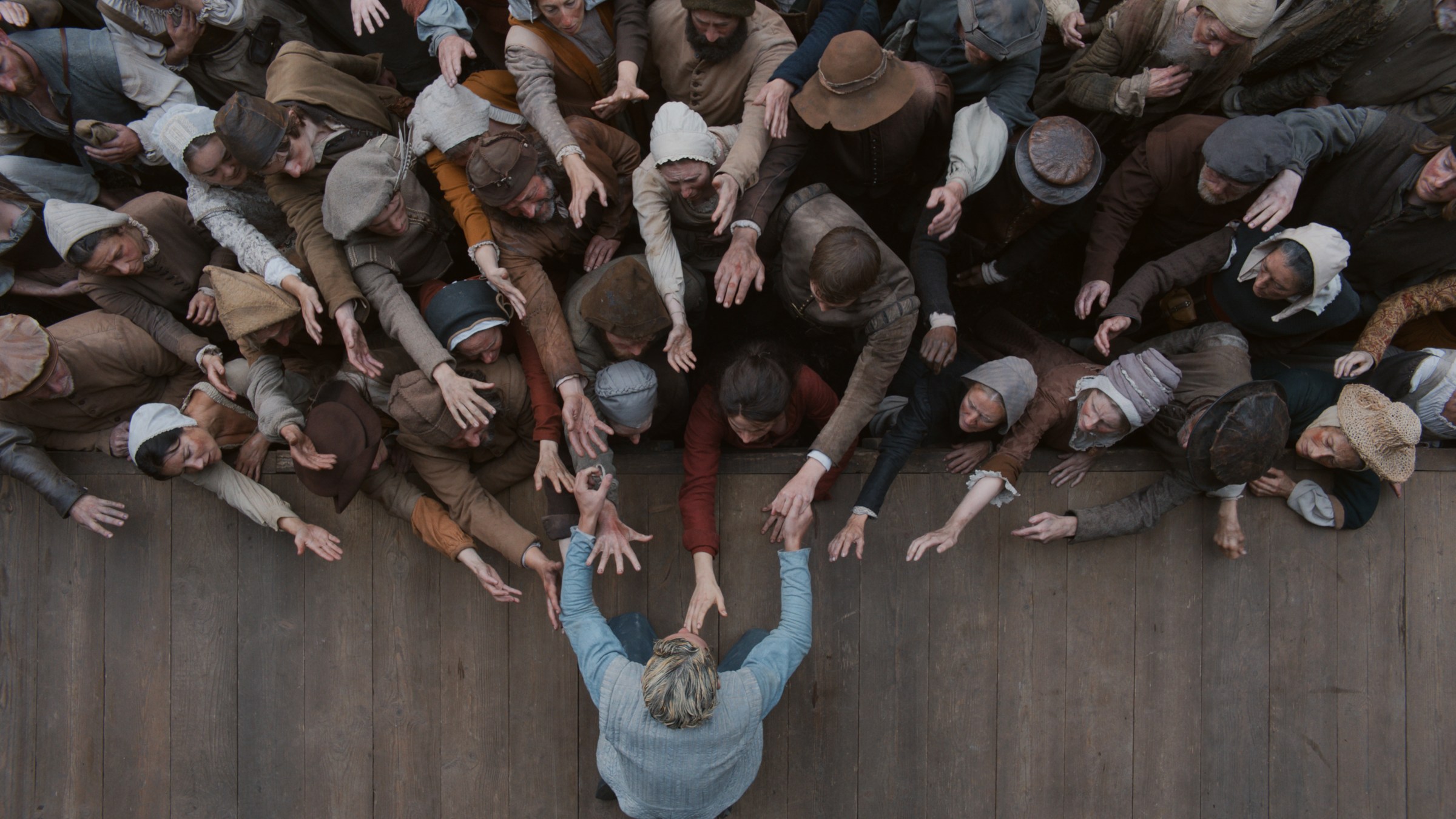 Seen from above, a blond man in blue reaches off the edge of a stage to a crowded audience, which is reaching back towards him.