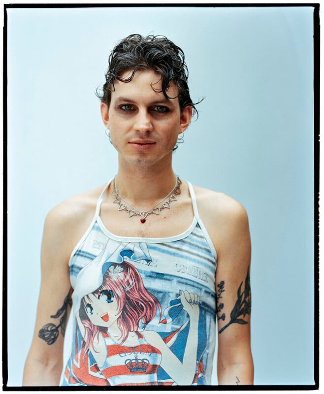 A person with short curly hair, wearing eyeliner and a necklace, stands against a light blue background. They have tattoos on both arms and are wearing a tank top featuring an anime-style character.