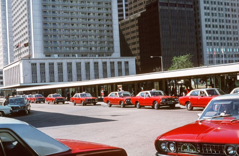A row of red taxis waits at a taxi stand in front of tall office buildings in an urban area; some drivers are visible inside the cars.
