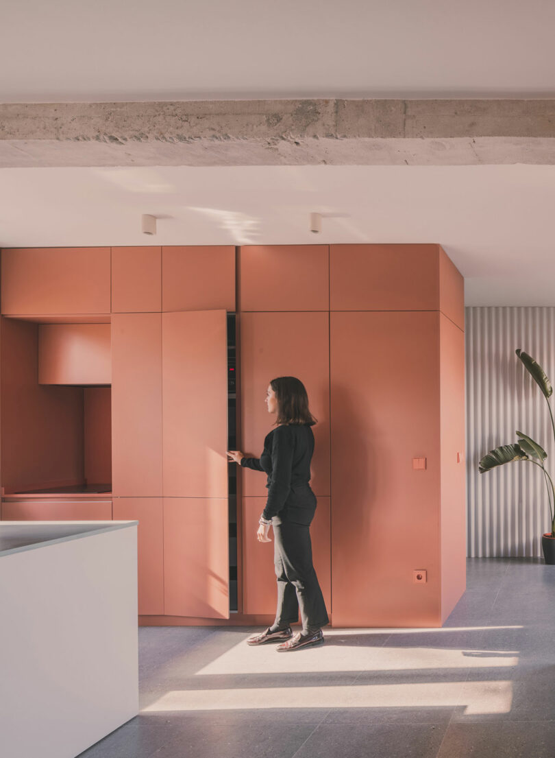 A person in black clothing opens a tall cabinet in a modern kitchen with matte orange cabinetry and minimalist design.