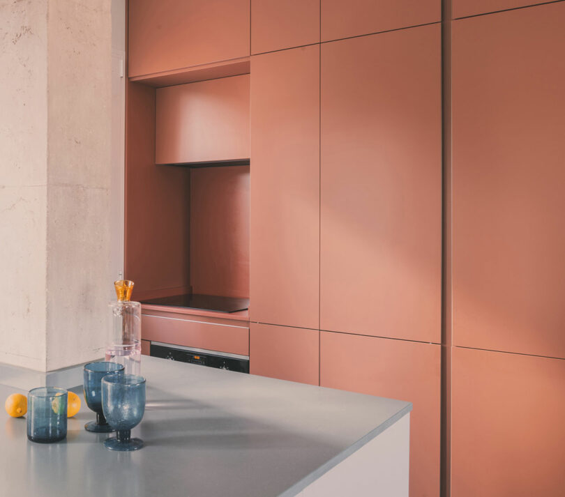 A modern kitchen with matte pink cabinets, a gray countertop, three blue glasses, a glass decanter with an orange stopper, and two lemons on the counter.