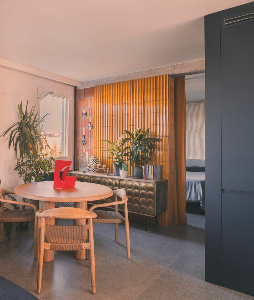 Modern dining area with a round wooden table, four chairs, potted plants, a red sculpture, and a retro sideboard. A yellow slatted divider separates the space from a bedroom.