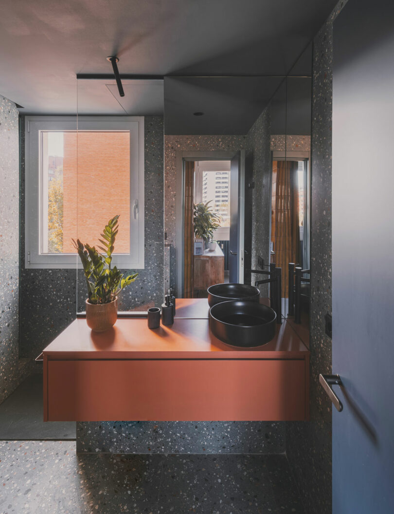 A modern bathroom with terrazzo walls and floor, an orange floating vanity, black dual sinks, a potted plant, large mirrors, and a window letting in natural light.
