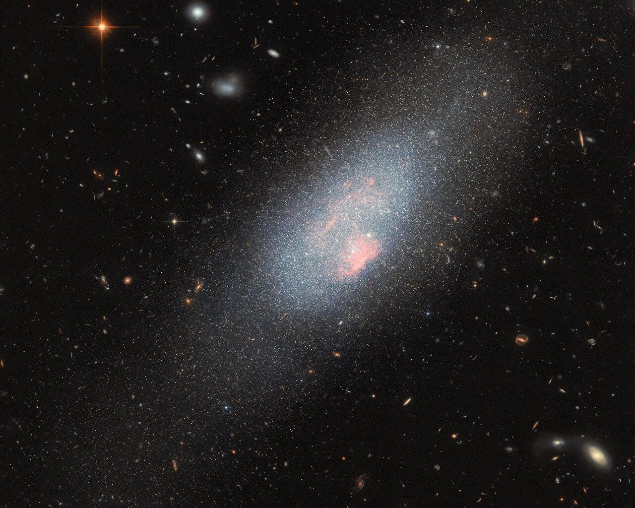 A pale blue dwarf galaxy on the black backdrop of space with some faraway galaxies. The galaxy itself resembles a fuzzy cloud of tightly packed stars, with a broad halo of stars dispersed around it. Spread across the galaxy’s core are several small, glowing patches of gas where there is a concentration of very hot stars.
