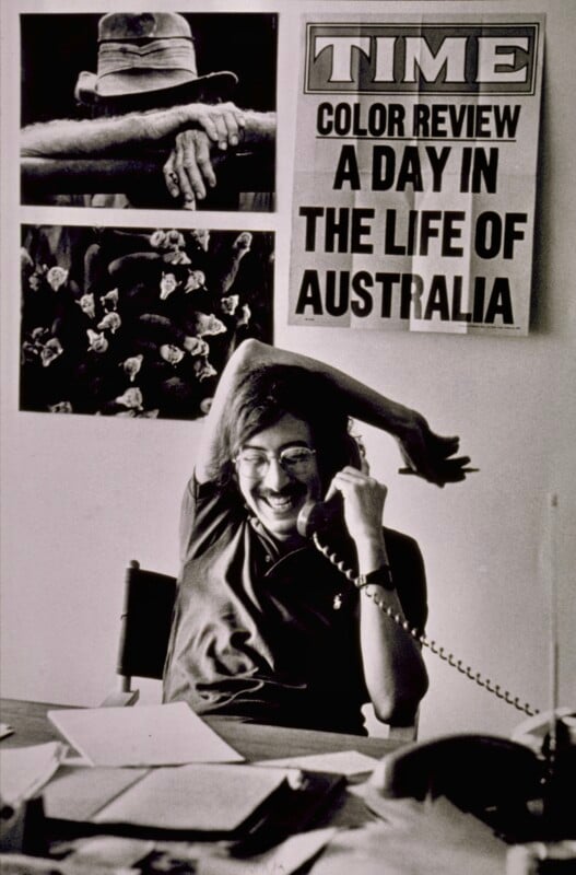 A person smiles while talking on a corded phone at a desk covered with papers. Behind them are two black-and-white photos and a TIME magazine poster reading “A Day in the Life of Australia.”.