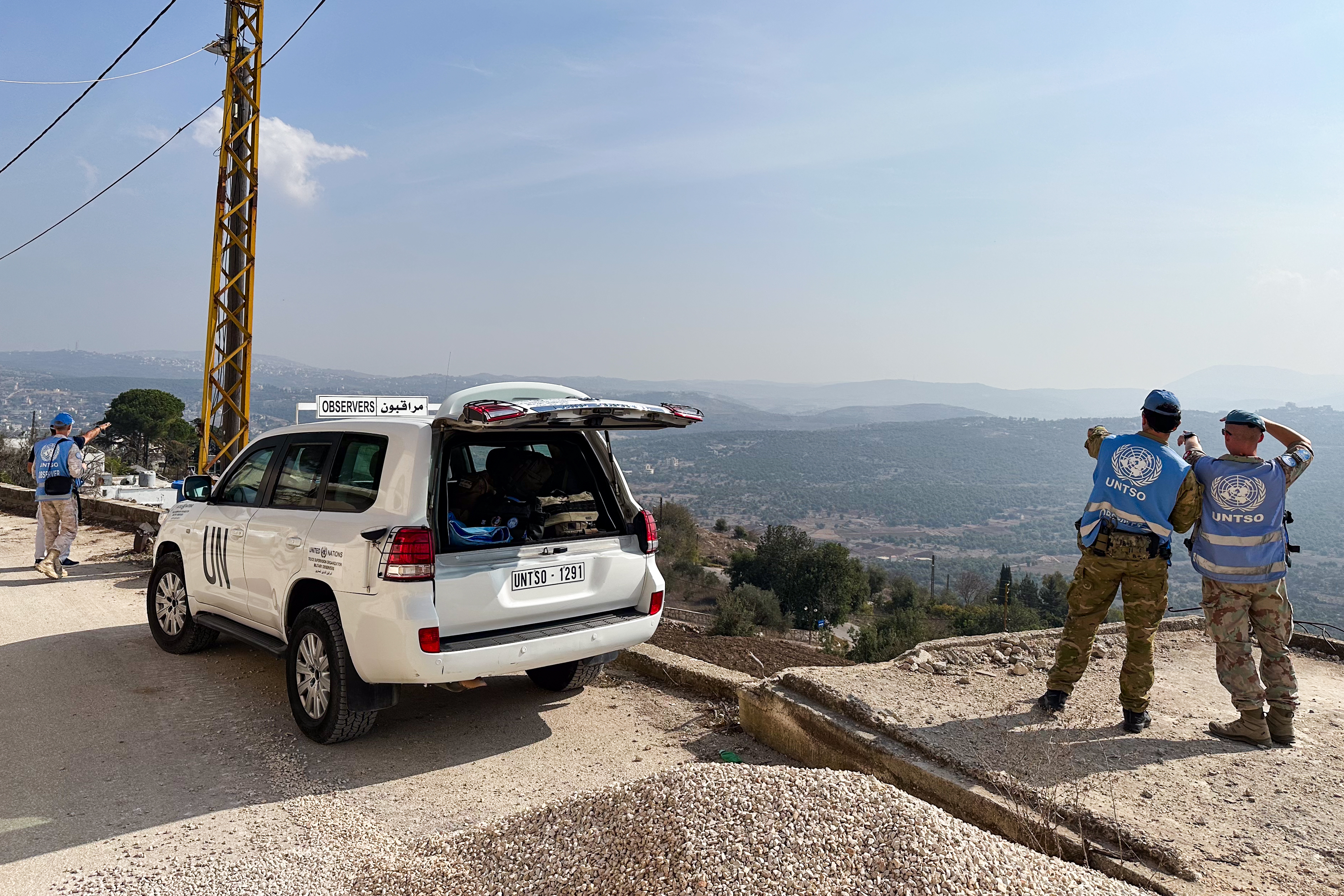 People in blue waistcoats overlook a landscape, a white car next to them