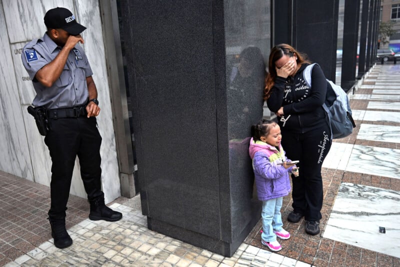 A security guard watches as a woman covers her face and stands next to a young girl crying against a building on a city sidewalk. The scene appears tense and emotional.