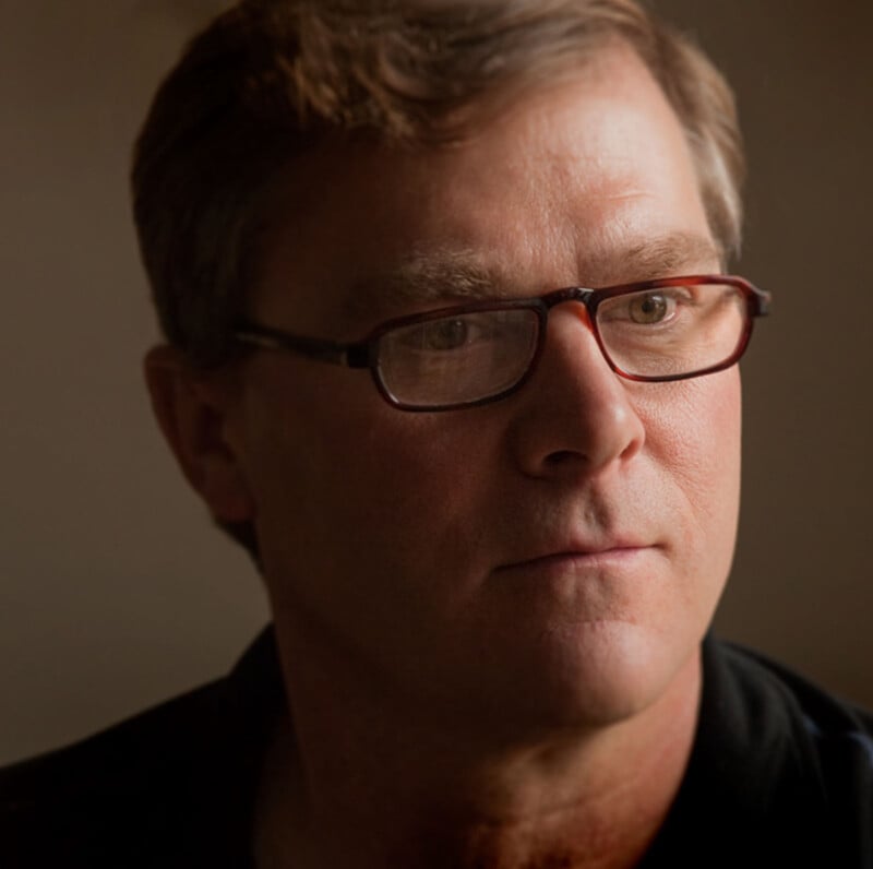 A man with short brown hair and glasses looks thoughtfully into the distance. He is wearing a dark shirt and is lit by soft, natural light against a neutral background.