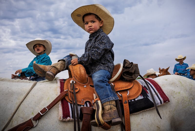 A young boy in a cowboy hat and boots sits on a saddled horse, looking at the camera. Another child in western clothing and hat is nearby, with a cloudy sky in the background.